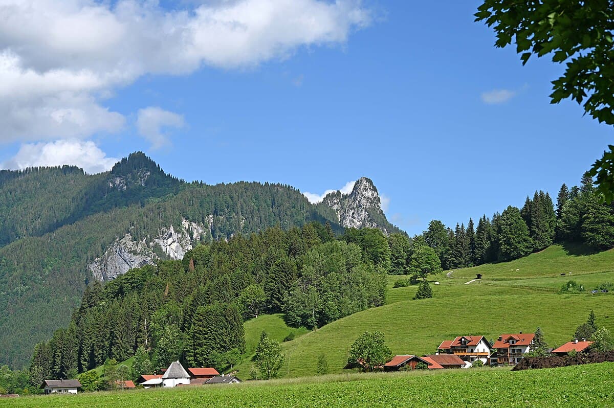 Ettal, im Hintergrund die Falkenwand, der vordere Rappenkopf in den Ammergauer Alpen und der markante Felsgipfel Kofel über dem Eingang ins Graswangtal bei Oberammergau. Nicht allzuweit ist auch das Ettaler Manndl, ein durchaus ähnlicher Gipfel ebenfalls oberhalb von Ettal, der schon öfter mal verwechselt wurde.